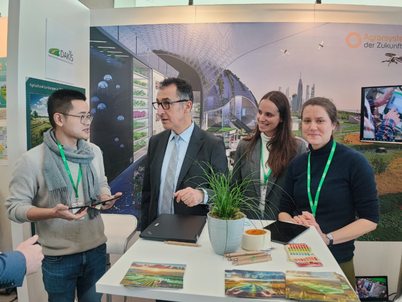 © DAKIS Ein Messestand, an dem vier Personen um einen weißen Tisch herumstehen. Im Hintergrund sind Grafiken und Fotos zum Thema Landwirtschaft zu sehen.