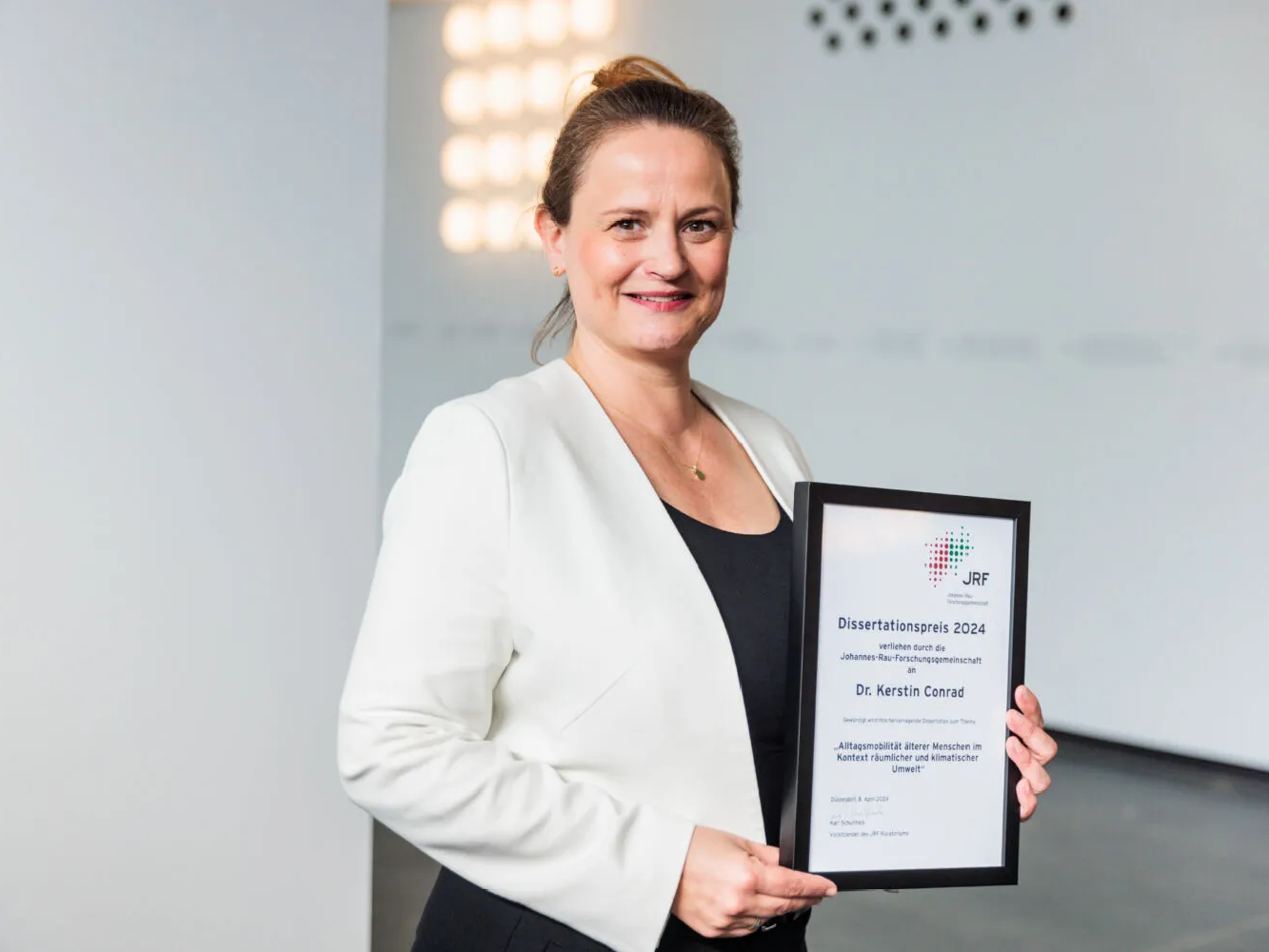 © JRF e.V. Kerstin Conrad hält in der Hand einen schwarzen Bilderrahmen mit der Auszeichnung.