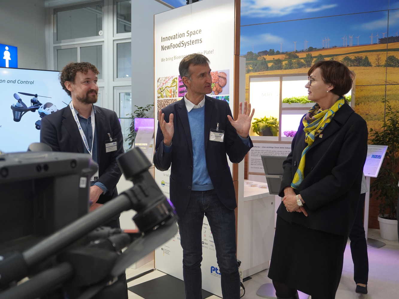 © Jette Behrend 3 Personen stehen nebeneinander. Die Person in der Mitte spricht mit der Person rechts, die Person ganz links hört zu. Im Hintergrund ist ein Messestand zum Thema Landwirtschaft zu sehen.
