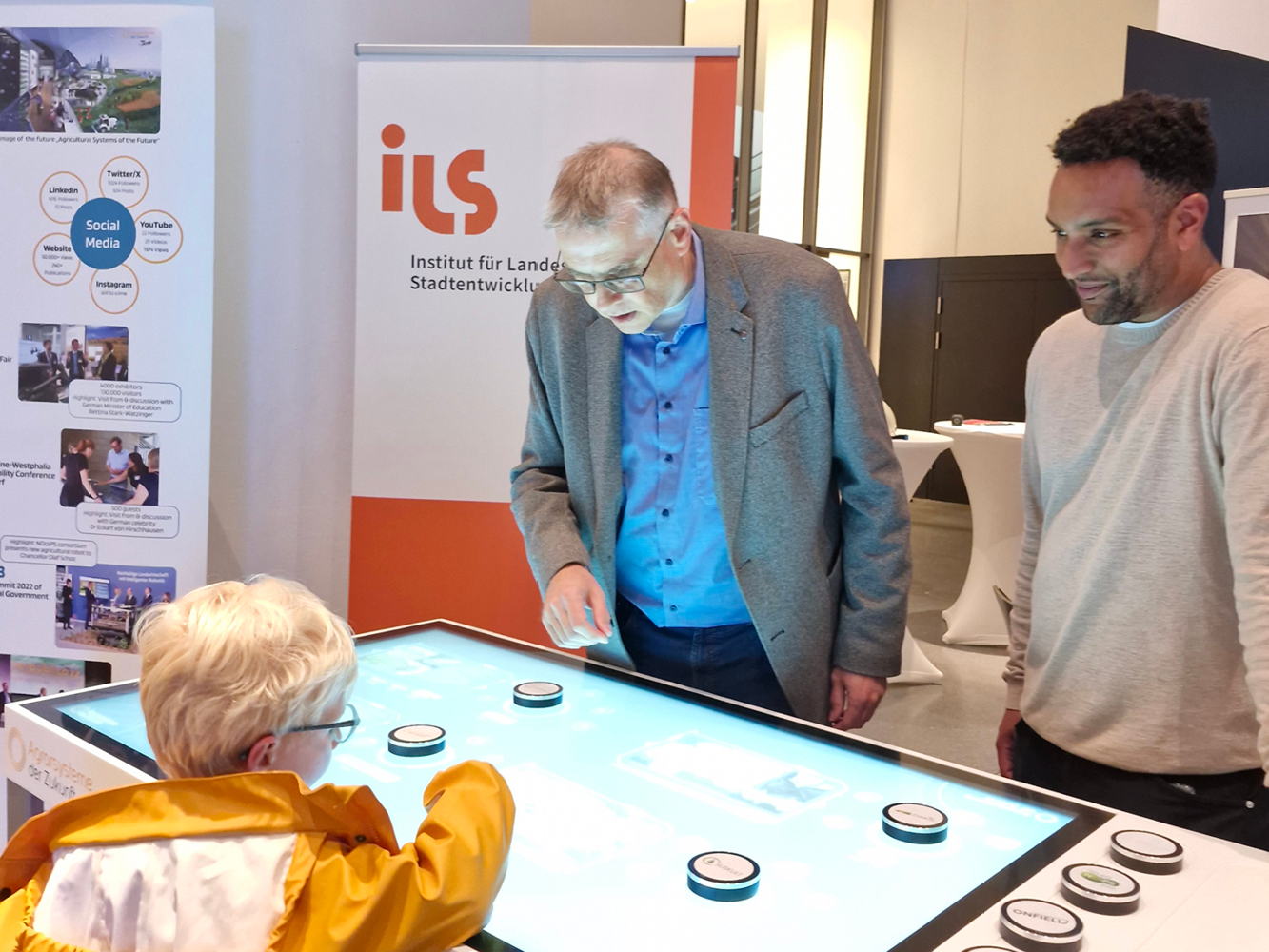 © ILS / von Bischopink Zwei Erwachsene und ein Kind stehen um einen Multitouch-Tisch herum. Das Kind zeigt auf einen Bereich auf dem Tisch und die Erwachsenen erklären und lächeln.