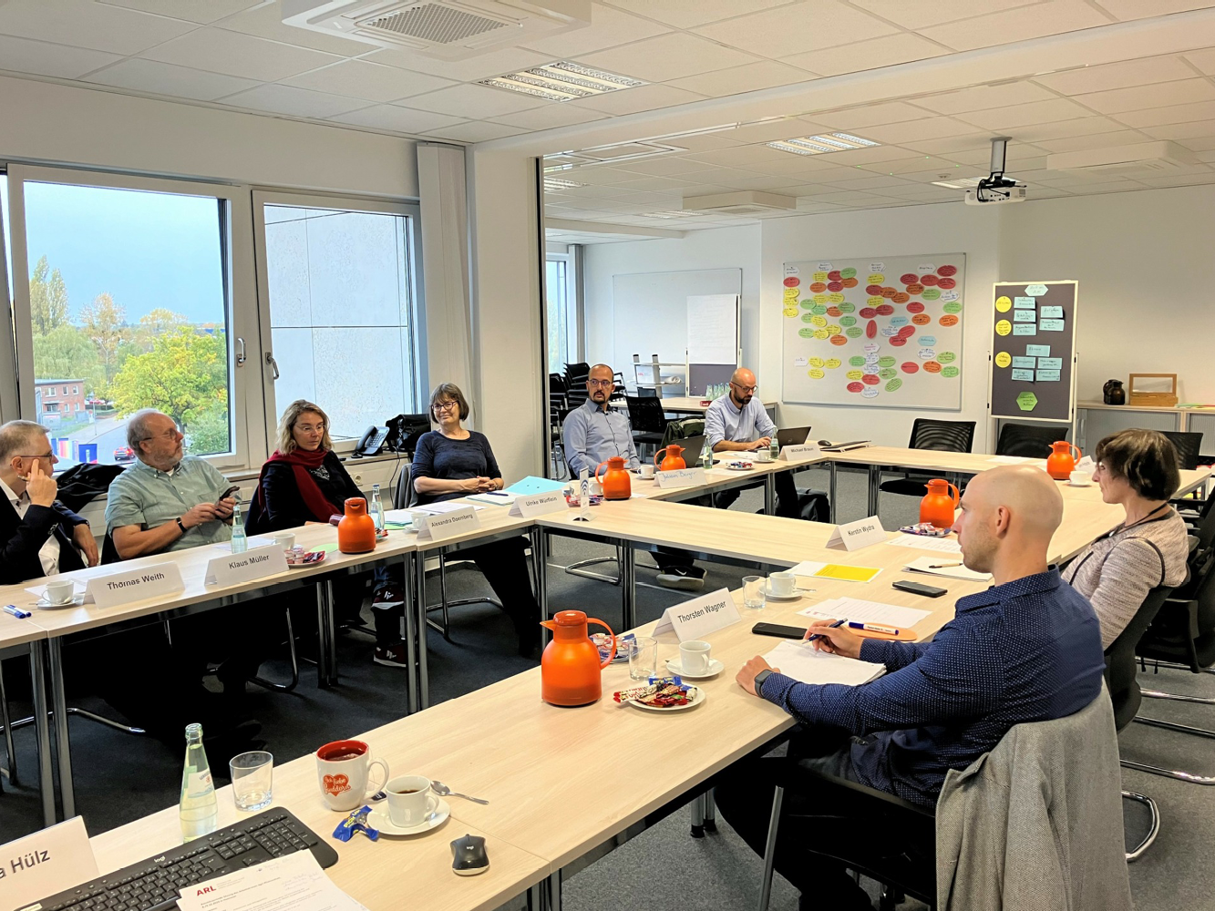 © ARL 8 Personen sitzen an Tischen, die zu einem Rechteck aufgestellt sind, so dass sich die Personen anschauen können. Im Hintergrund sind Whiteboards zu sehen.