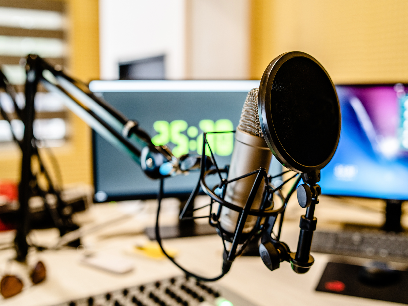 © AdobeStock_270487782 Im Vordergrund ist ein Mikrofon an einer Aufhängung zu sehen, im Hintergrund Monitore und ein Mischpult.