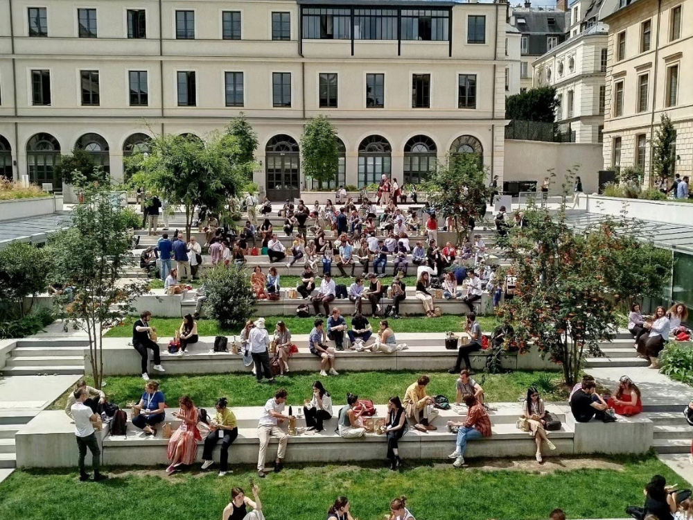 © Stefano Cozzolino / ILS Ca. 100 Personen sitzen vor einem Gebäude in der Sonne. Sie sitzen auf Mauern, die hintereinander in größerem Abstand vor dem Gebäude als Sitzmöglichkeiten installiert wurden.