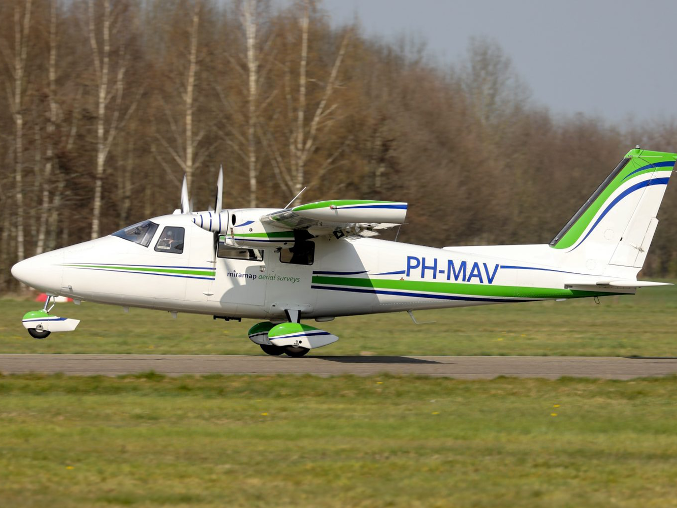 © Miramap Aerial Surveys Ein weißes Propellerflugzeug mit grünen und blauen Linien auf einer Startbahn.