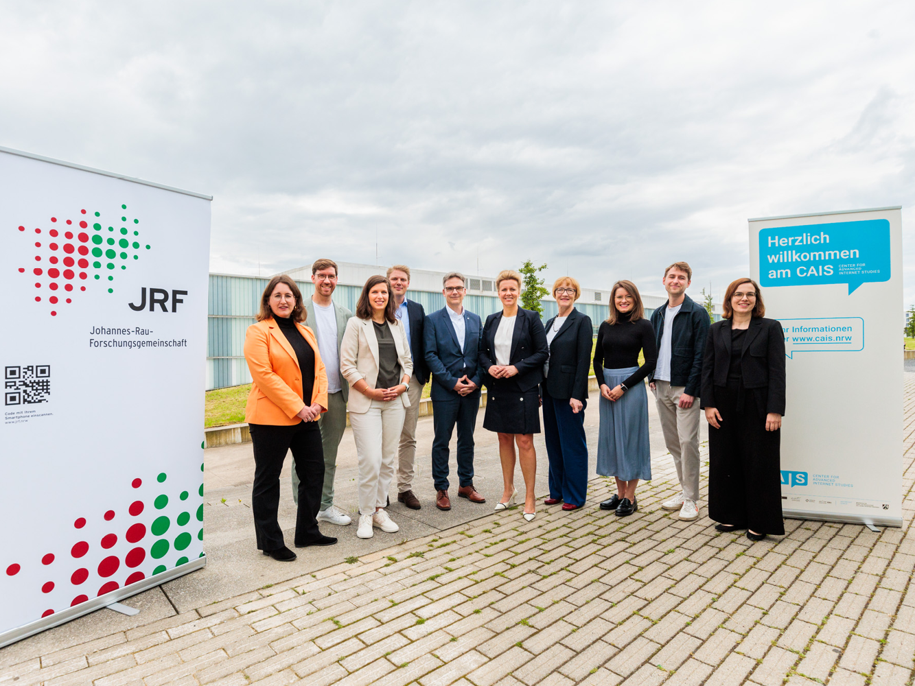 © JRF e.V. 10 Personen stehen nebeneinander und lächeln in die Kamera, links und rechts von ihnen jeweils ein Roll-up mit Logos der an der Veranstaltung beteiligten Organisationen.