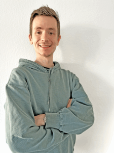 Gideon_Weier_klein junger Mann in pastellgrüner Kapuzenjacke lächelt freundlich in die Kamera. Er hat die Arme vor der BRust locker gekreuzt.