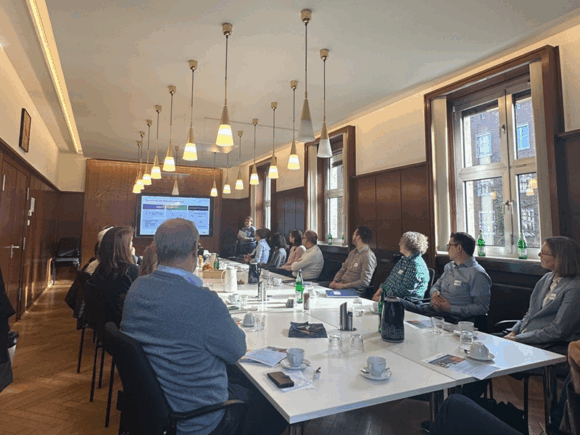 Personen sitzen an einem langen Tisch mit Blick nach vorne zur Person, die die Runde begrüßt.