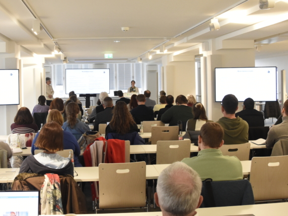 Blick in einen Veranstaltungsraum mit Tischenund Stühlen, vorne leuchten drei große Bildschirme mit Präsentationen und Fotos.