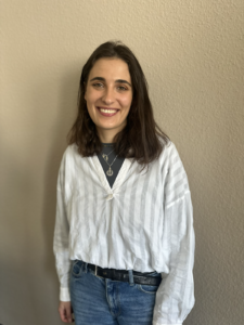 Junge Frau lächelt in die Kamera, steht vor einer beigefarbenen Wand und tragt Jeans und eine weiße Bluse.