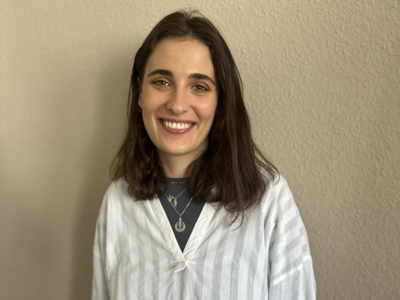 Junge Frau steht vor einer beigen Wand und lächelt in die Kamera