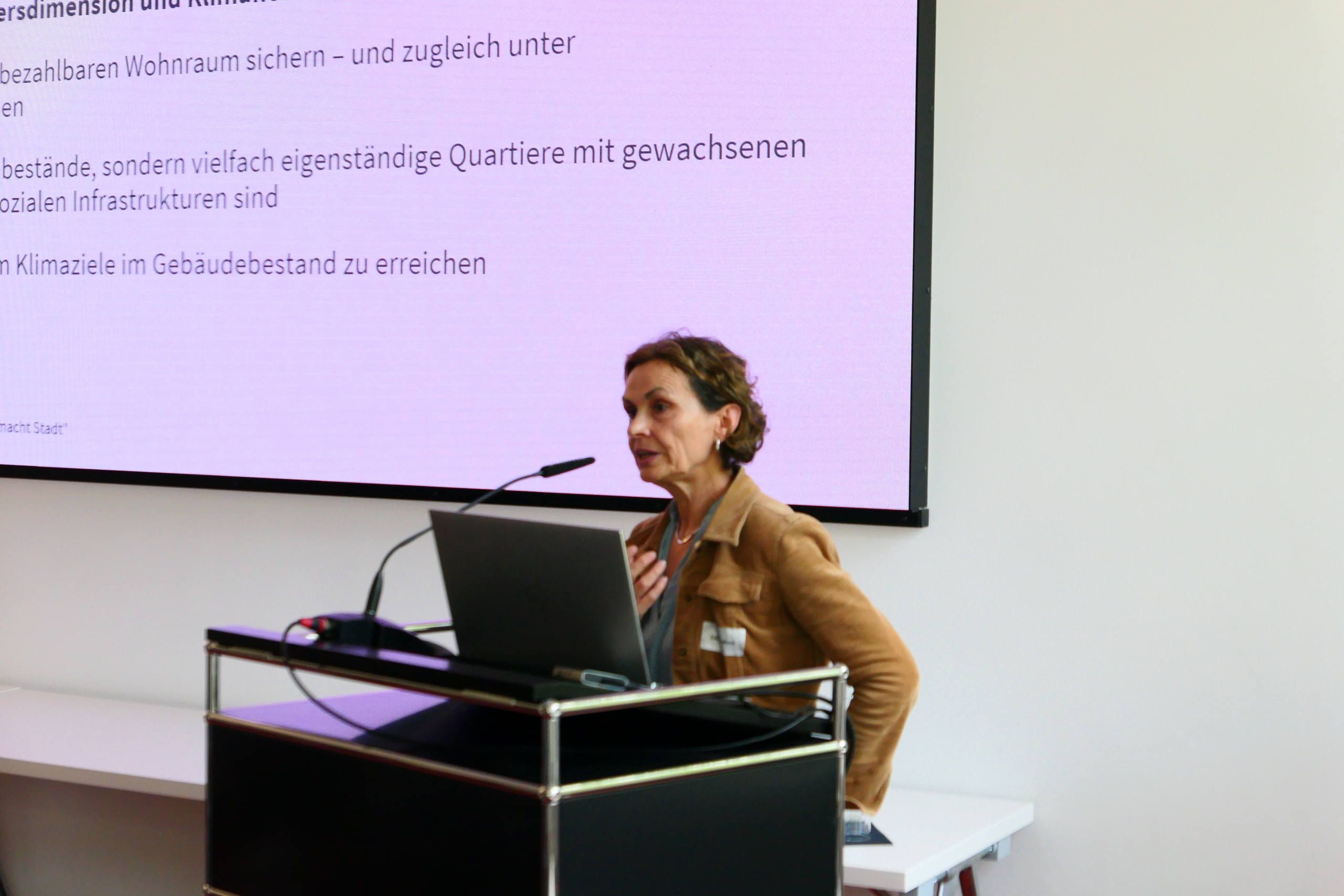 P1030648 Dr. Sabine Weck steht an einem Redepult und spricht in ein Mikrofon. Im Hintergrund ist ihre Präsentation zu sehen.