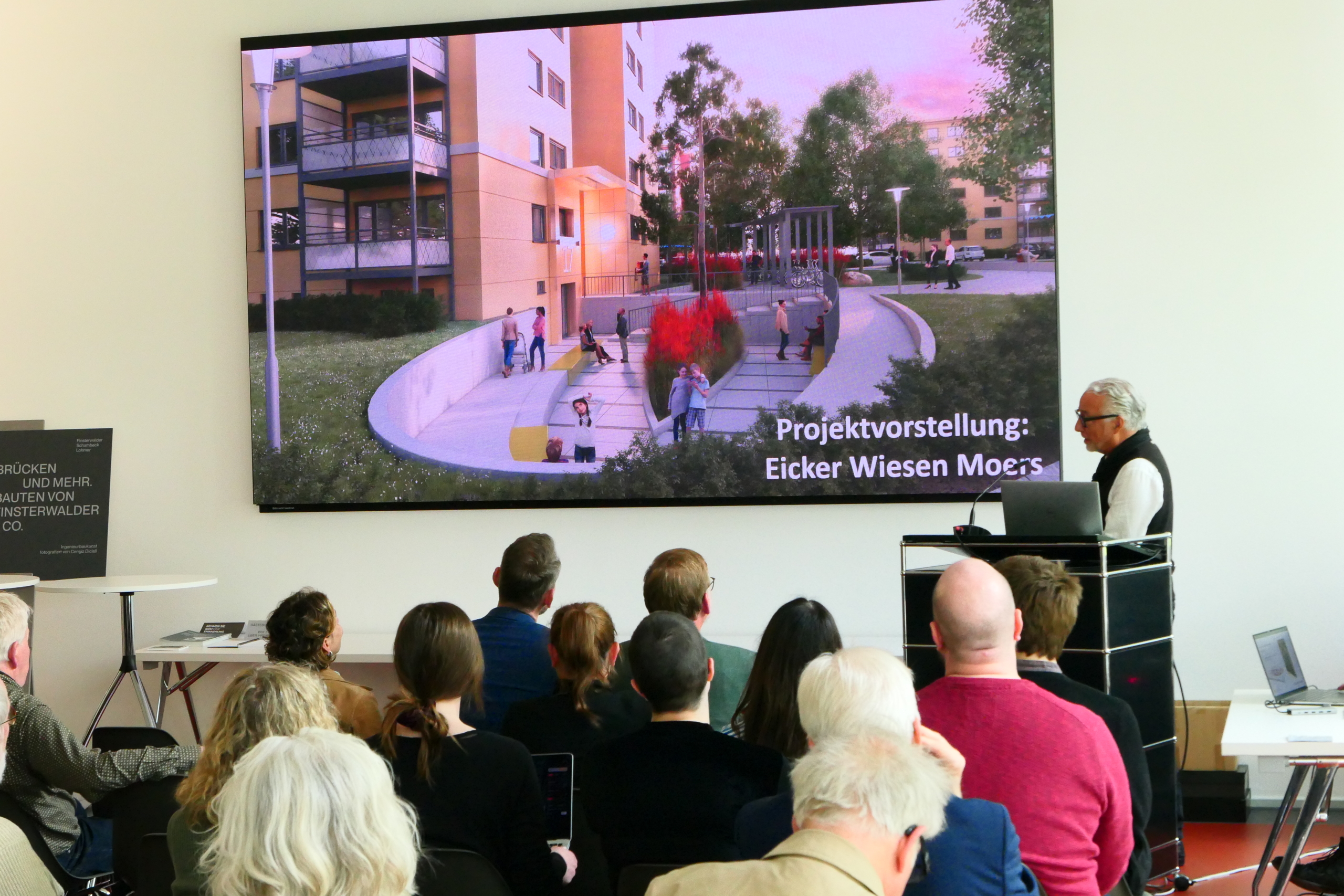 P1030677 Ein Publikum auf STühlen schaut nach vorne auf eine Präsentation auf einem großen Bildschirm.