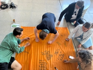 Fünd Personen sitzen auf dem Boden um ein Tuch in orange und schreiben mit Edding auf das Tuch.