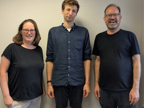 3 Personen, vor einer hellen Wand, lächeln in die Kamera