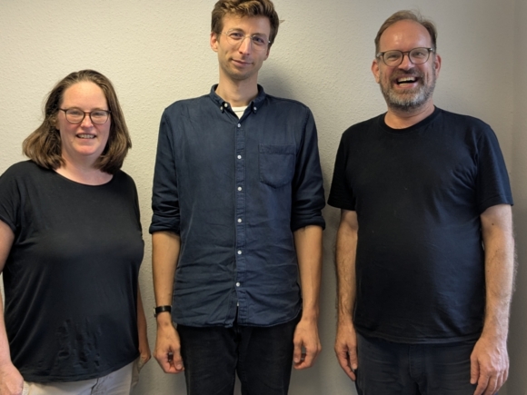 3 Personen, vor einer hellen Wand, lächeln in die Kamera